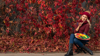 The red maple leaves a woman artist paints on the wall of leaves with live colors in blue jeans and a burgundy sweater. Smiling with a lovely smile sits
