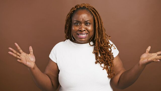 Emotional african american woman gesturing hands in disbelief, laughing nervously, brown studio background