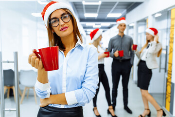 young business woman in Santa Claus hat, holding a red mug, with a group of colleagues in the...