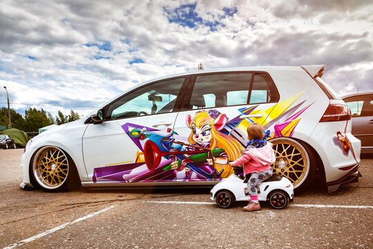 Moscow, Russia: July 06, 2019: Baby sits on a toy vehicle near tuned by low suspension and custom colored wheels. White golf 7 with aerography on which painted Chip and Dale characters.