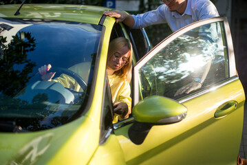 Naklejka premium A woman with blonde hair is sitting in a yellow car and a man in a shirt is standing near. Auto business, car sale, consumerism and people concept - happy woman with car dealer. 