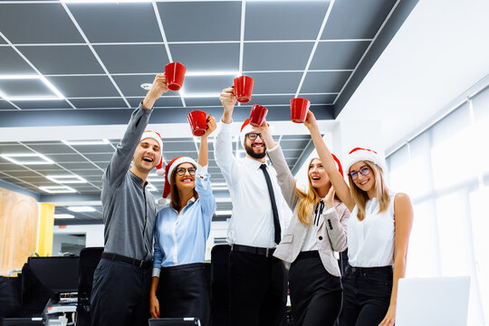 Group of successful businessmen in Santa Claus hats celebrating a holiday in a modern office. Young people drink a drink and have fun. Happy New Year and Merry Christmas