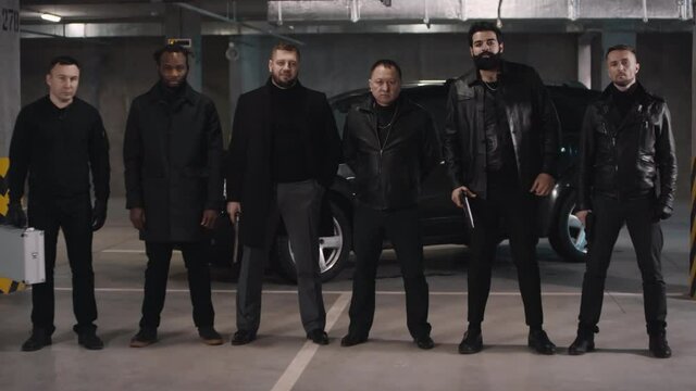 Full portrait of sex multi-ethnic brutal criminals standing in row wearing black clothes holding guns and metal briefcase and black car in background at underground parking