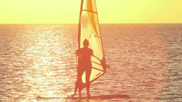 Silhouette of woman sailing windsurf board along tranquil ocean at bright back sunset light at tropical resort backside view