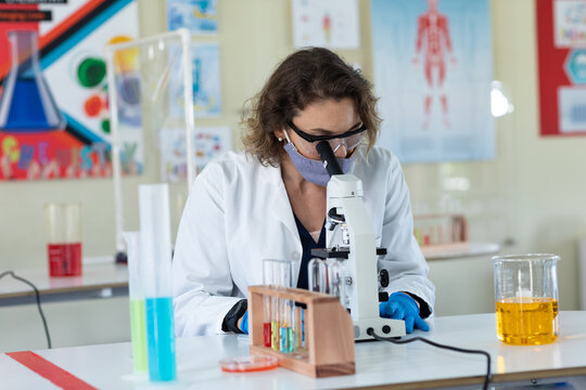 Female teacher wearing face mask and protective glasses using microscope in laboratory - Powered by Adobe