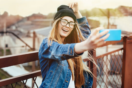 Young sexy blonde hipster woman posing for selfie and laughing. Wearing jeans jacket, hipster black hat and glasses. Lifestyle portrait bright with sun shine.