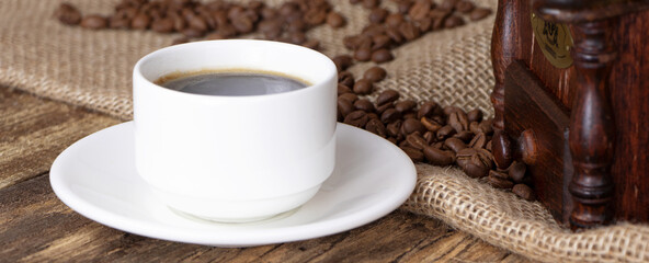 A white cup of coffee on a wooden background. Banner. A coffee grinder and coffee beens on a linen fabric.