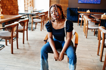Lovely african american woman with dreadlocks in blue stylish jeans at cafe. Beautiful cool fashionable black young girl indoor.