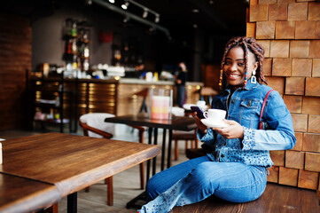 Lovely african american woman with dreadlocks in blue stylish jeans jacket at cafe hold mobile phone. Beautiful cool fashionable black young girl indoor drink coffee.