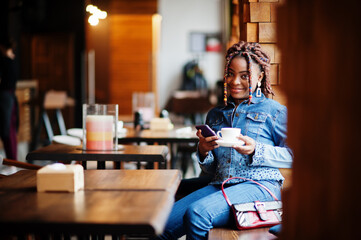 Fototapeta na wymiar Lovely african american woman with dreadlocks in blue stylish jeans jacket at cafe hold mobile phone. Beautiful cool fashionable black young girl indoor drink coffee.