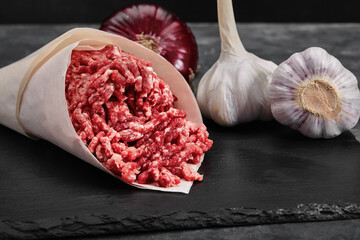 Minced meat on paper with seasoning and fresh garlic on black background, side view
