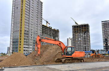 View of a large construction site where earthmoving equipment. Excavator and wheel loader digs the ground to lay pipes and a new road. Tower cranes are building tall residential buildings.
