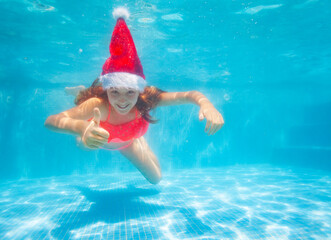 Happy girl with thumb up gesture dive and swim underwater wearing Santa Claus hat in the pool