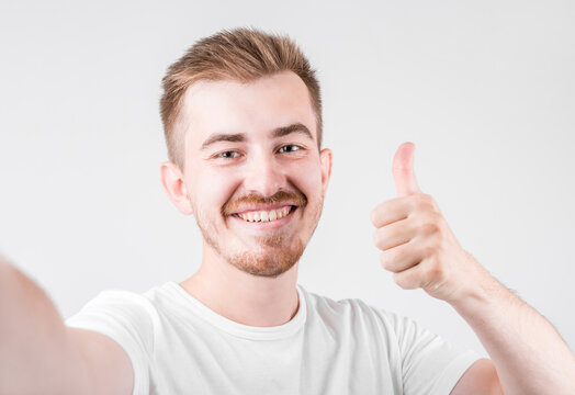 Photo of handsome man in casual t-shirt , smiling on camera with thumb up while taking selfie isolated over white background