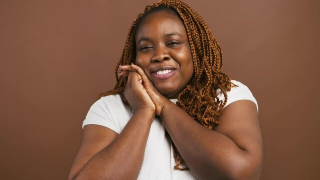 Grateful black woman clasping hands and smiling, touched by complement, brown studio background