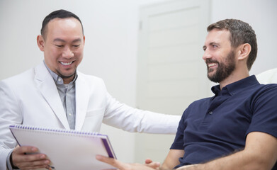 Fototapeta premium Young handsome man in beauty salon consults with beautician on anti-aging treatments. Doctor writing notes on clipboard and explain procedures to the patient