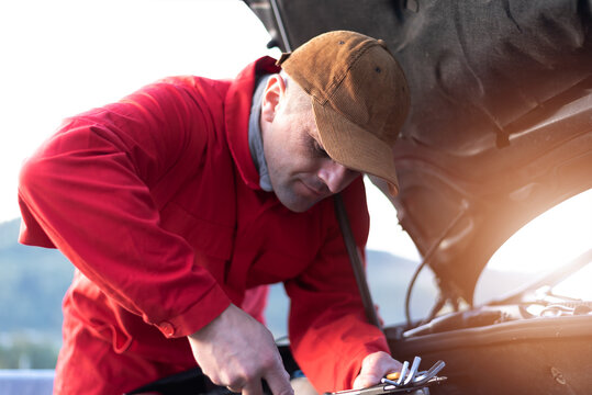 Handsome auto mechanic or road assistance worker in uniform repairing engine of the broken car on the road