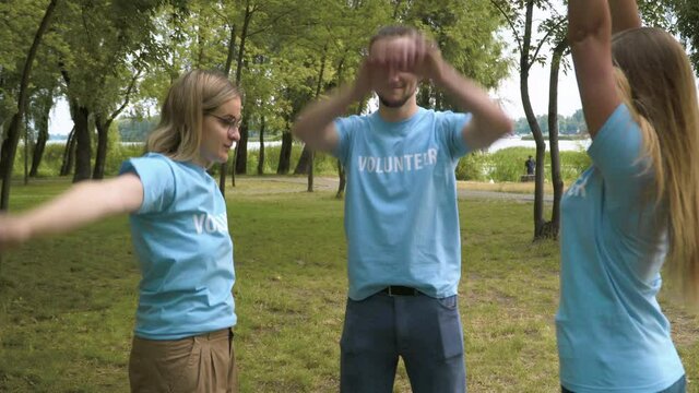 Satisfied volunteers stacking hands standing on clear meadow in summer park. Portrait of young Caucasian man and women rejoicing cleanup. Environmental and ecological protection.