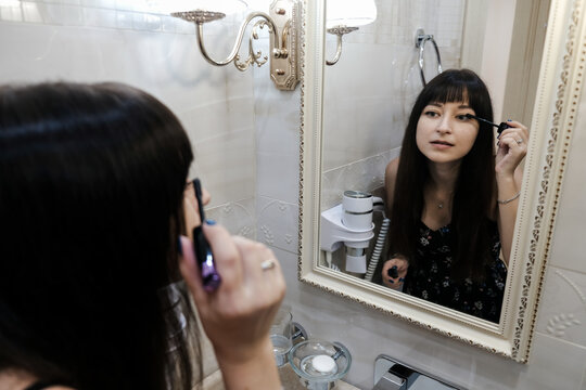Young woman applying mascara in front of a mirror. Girl getting ready with her makeup in a hotel bathroom. Pretty female serious face while brushing eyelashes, going out for the evening with friends.