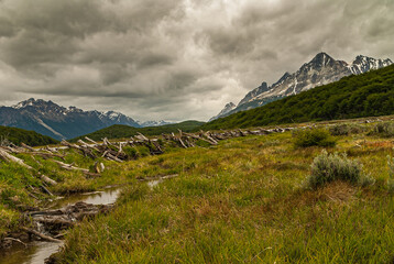 Ushuaia, Tierra del Fuego, Argentina - December 13, 2008: Martial Mountains in Nature Reserve. Landscape with long dam, brook in grassland, snowy mountains in back under gray cloudscape.