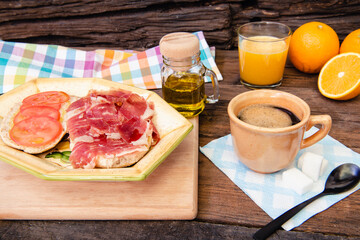 Desayuno típico español, una taza de café, pan con jamón y rodajas de tomate, una botella de aceite de oliva, naranjas y un vaso de jugo, sobre fondo rústico