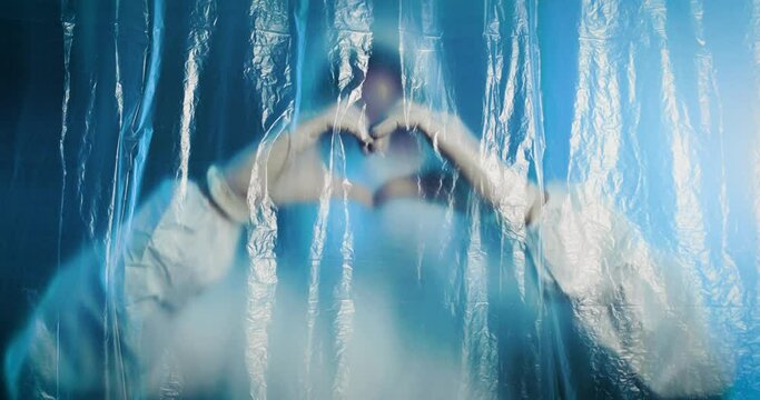 Medico o infermiere con tuta covid fa il simbolo del cuore dietro a un telo di plastica