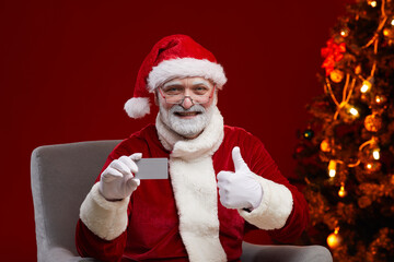 Senior smiling man in Santa's costume holding business card and showing thumb up to the camera sitting on armchair