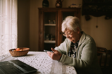 Senior caucasian female with glasses while sitting at a table and writing messages on a mobile phone during quarantine COVID - 19 coronavirus