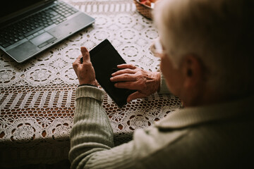 Fototapeta na wymiar Close up of a caucasian senior woman's hand while typing on a tablet, while in front of her on the table is a laptop during quarantine COVID - 19 coronavirus