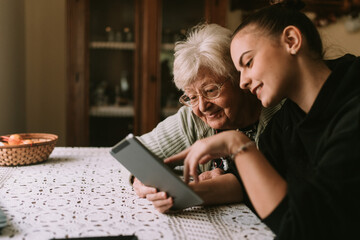 Smiling caucasian grandmother and beautiful granddaughter in the kitchen looking at the tablet. The...