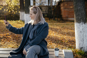 Beautiful young happy 30-year-old woman takes a selfie with her smartphone on a bench in an autumn Park. The concept of the season, technology and people .