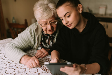 The senior caucasian  grandmother and her beautiful teenage granddaughter look at the pictures on the tablet in the room
