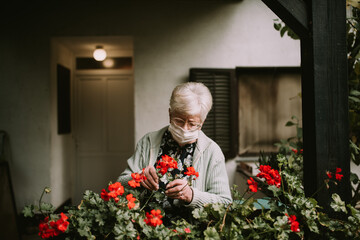 Cute caucasian grandmother with glasses and a face mask arranges flowers in her garden during the quarantine of the epidemic COVID - 19. Protection of the elderly in a coronavirus pandemic