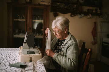 Side view of a retired caucasian senior woman with eyeglasses holding a white cloth in her hand and sewing on a sewing machine in the living room