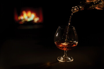 Woman pours cognac into a glass on the background of the fireplace.
