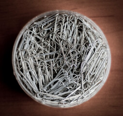 A heap of paperclips in a plastic container on a wooden table. A close-up with selective focus.