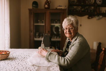 Portrait of a caucasian grandmother with retired glasses who sews a curtain on a sewing machine on...