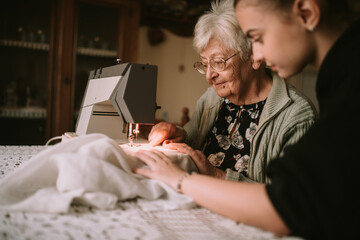 A teenage caucasian girl helps her senior grandmother to sew a dress on a sewing machine with a lamp