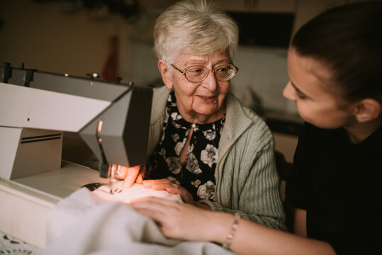Senior cute smiling caucasian grandmother and her teenage granddaughter talk while teaching her to sew clothes on a sewing machine