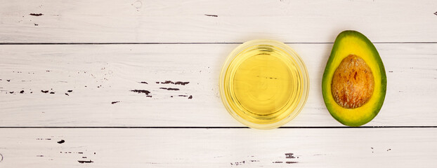 Avocado oil in a glass Cup and avocado cut with a stone on a white wooden background, top view, copyspace. Healthy food, vitamins, vegetarian food.