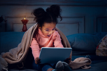 Happy african-american little girl during video call with laptop and home devices, looks delighted and happy. Talking to Santa before New Year's eve, her family, taking online preschool class.