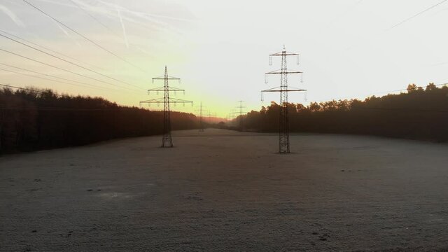 Aerial fly backwards low angle shot, revealing sunrise lighting high voltage electric poles amidst foggy forest strip
