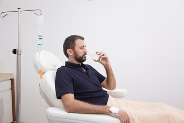 Sick, dehydrated or hangover patient man receiving vitamin IV infusion drip and drinking glass of...