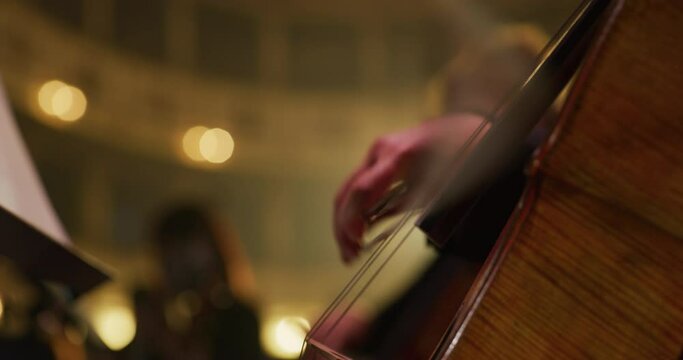 Cinematic close-up shot of female cellist is playing cello during musical concert on classic theatre stage with symphony orchestra performing on background.