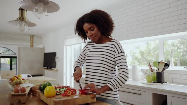 Happy young african woman cutting red capsicum in slices on wooden chopping board in modern kitchen