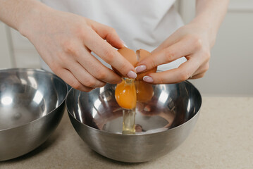 A close photo of the hands of a young woman who is breaking the organic farm egg above the stainless steel soup bowl in the kitchen.