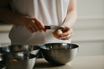 A dark photo of the hands of a young woman who is breaking the eggshell of the organic farm egg with the knife above the stainless steel soup bowl in the kitchen