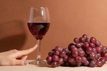 Tender woman finger touch the wineglass with red wine with branch of grapes background. Beige surface.
