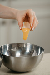 A close photo of the hand of a young woman who is pouring the yolk from the shell of the organic farm egg to the stainless steel soup bowl in a kitchen.