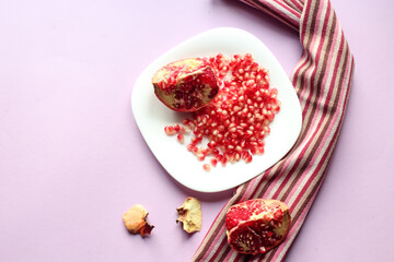 Cut pomegranate fruit with grains on a white plate, kitchen napkin, chrysanthemums on a purple background, top view-the concept of eating a healthy vegetable